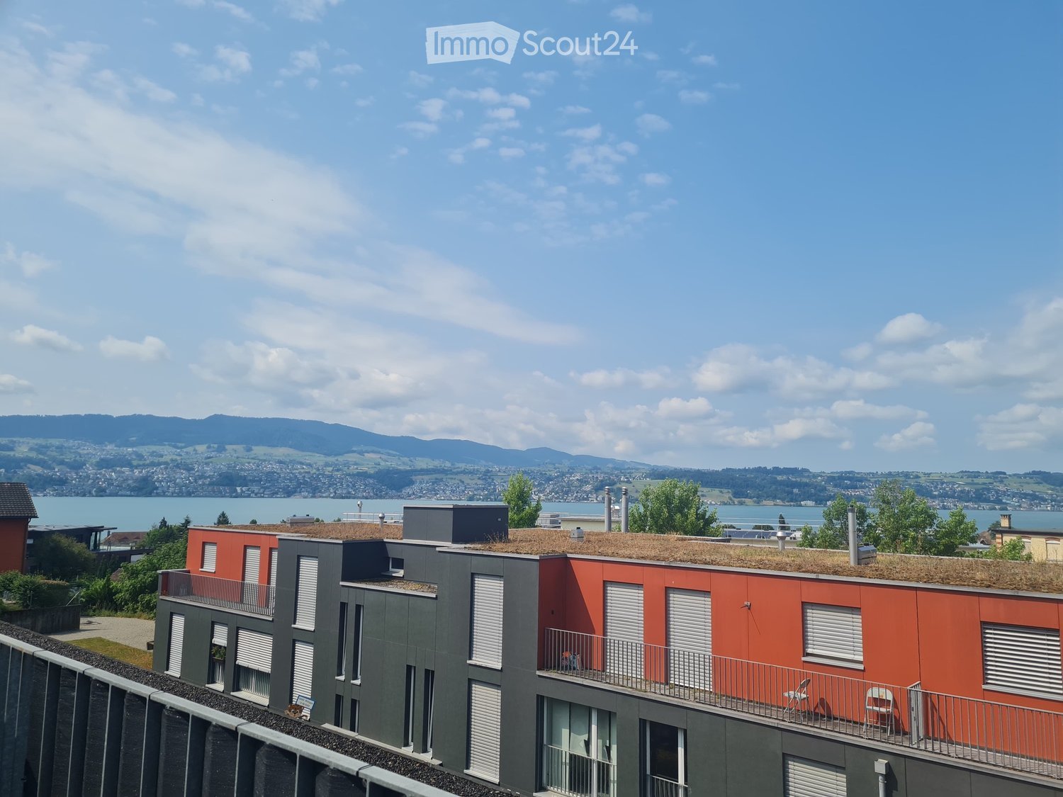 Modern multi-story building with red and grey exterior, featuring balconies, a green roof, and a scenic view of a lake and distant mountains.