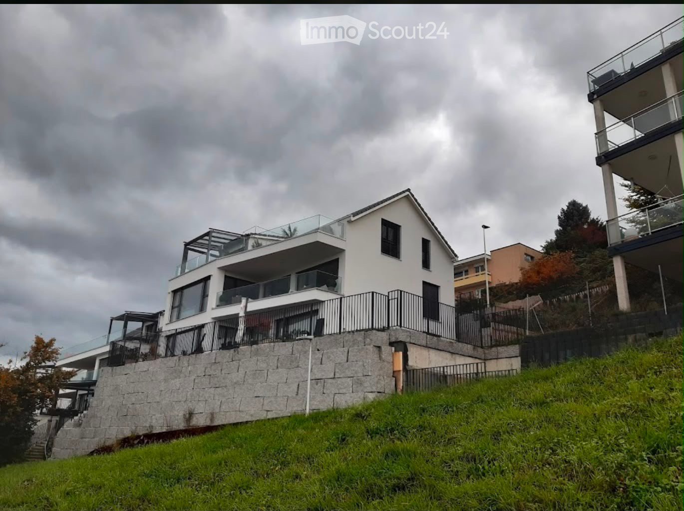 Modern two-story house with white exterior, large windows, and multiple balconies, situated on a raised ground floor with stone retaining wall and green lawn.