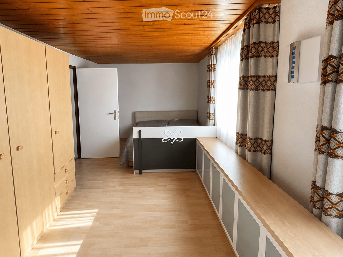 Cozy bedroom with wooden flooring, a bed with a headboard, a wardrobe, and patterned curtains on large windows.
