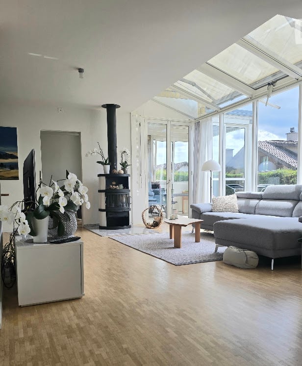 Bright living room with wooden flooring, large windows, a fireplace, a grey sectional sofa, coffee table, floor lamp, and a television on a stand.