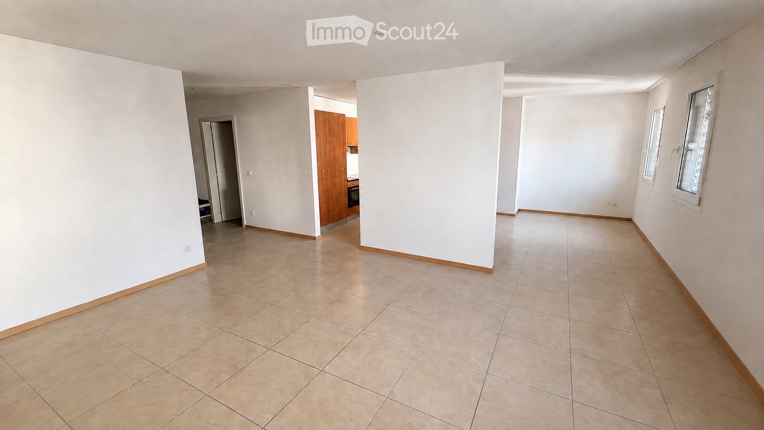 Empty room with tiled floor and white walls, featuring a doorway leading to another room with wooden cabinets and a small kitchen area.