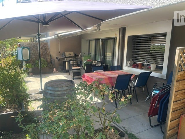 Outdoor patio area with a large umbrella, dining table with chairs, barbecue, and surrounding greenery.