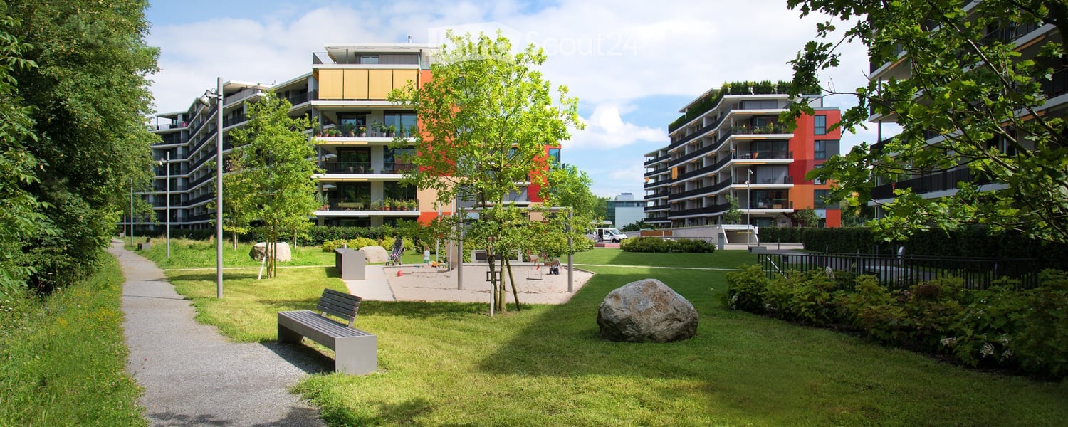 A well-maintained park area with a bench, trees, and a large rock, surrounded by modern apartment buildings.