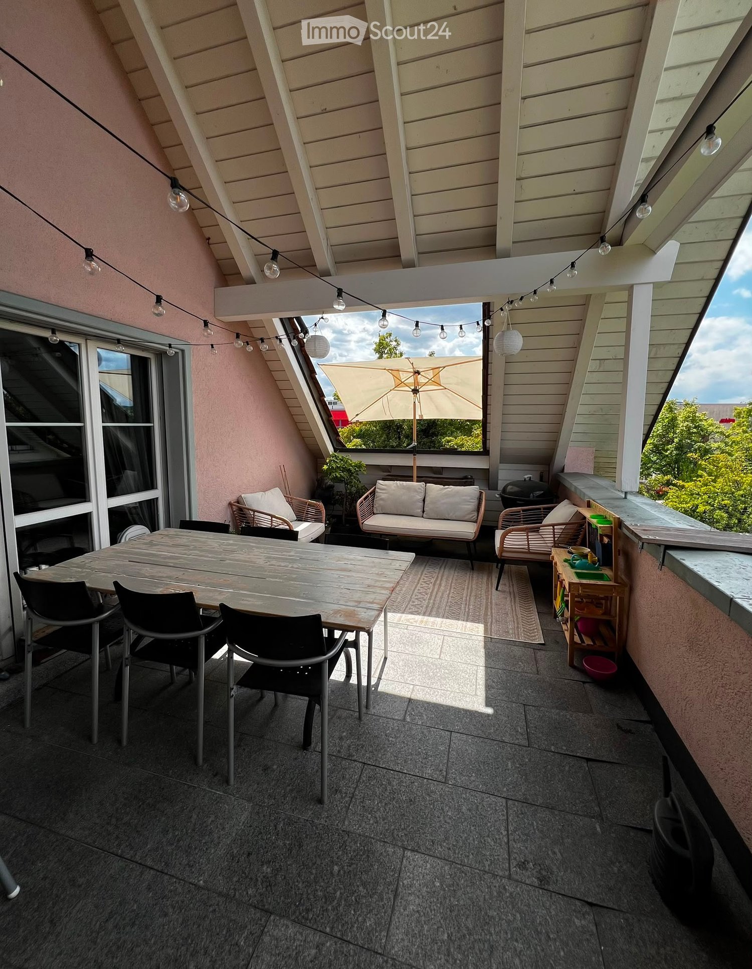 Balcony with wooden table, chairs, sofa, umbrella, and storage unit, overlooking a green area.
