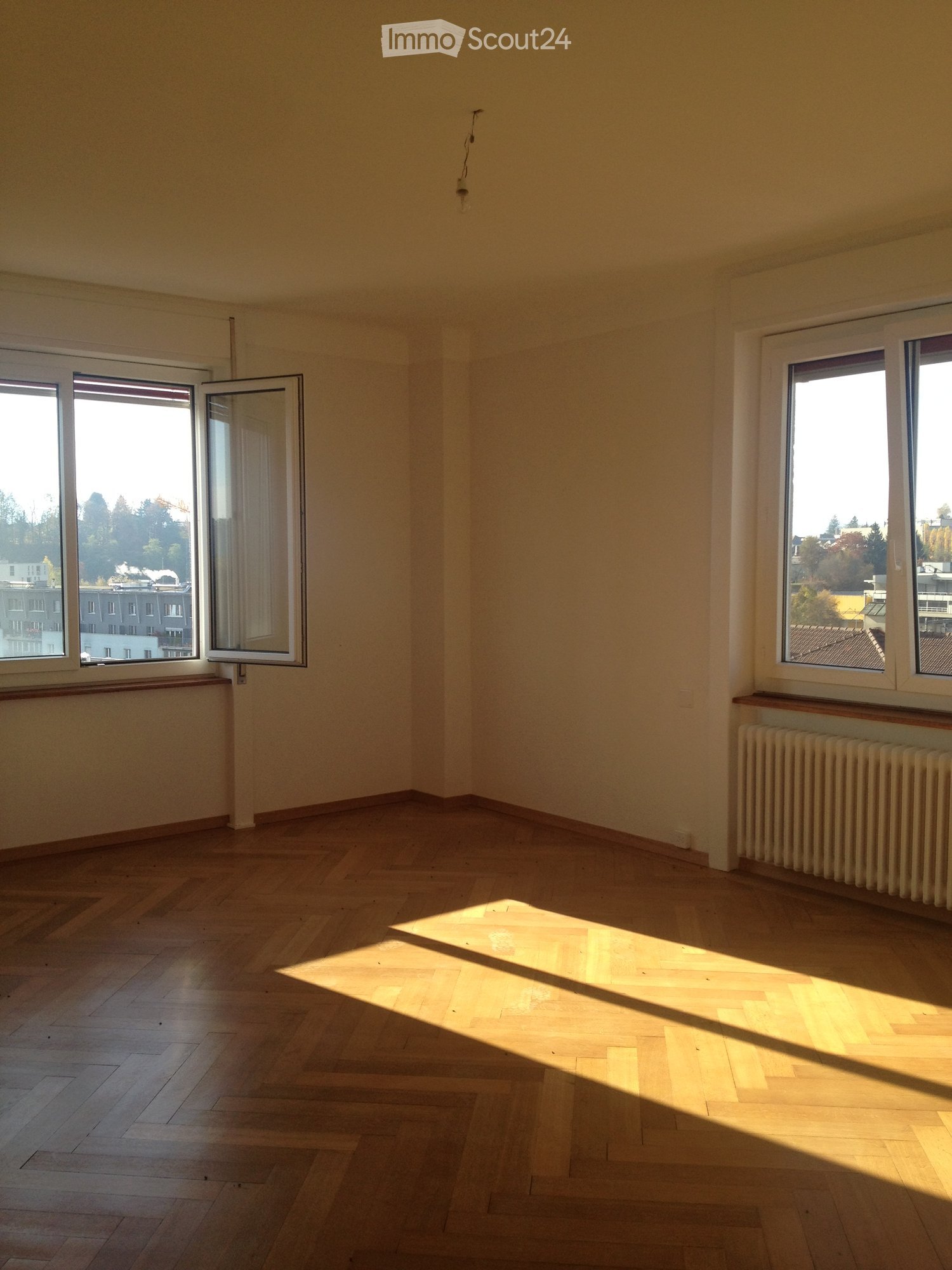Empty room, parquet floor, white walls, two windows, radiator, and outside view
