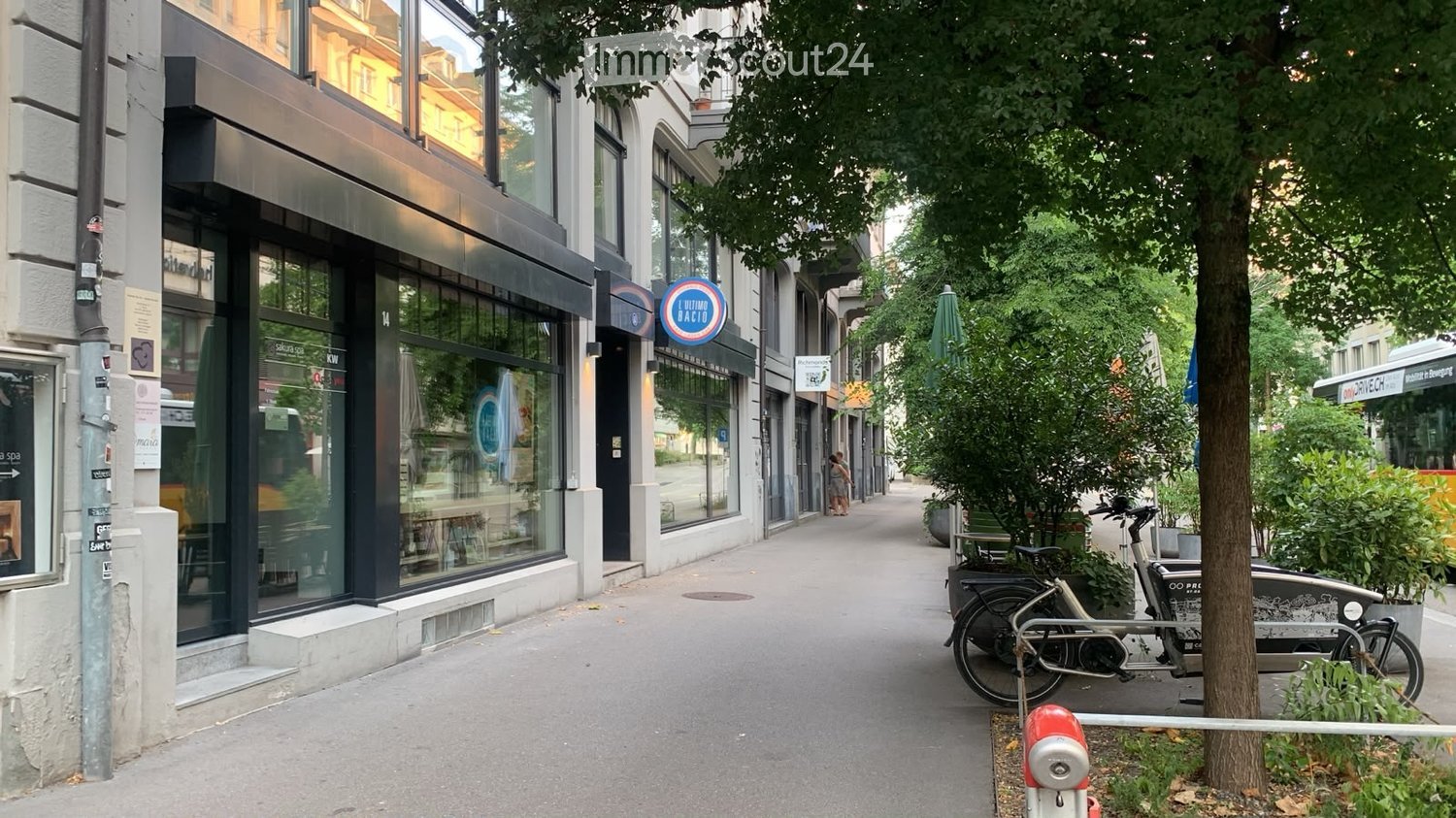 A modern urban street with a mix of commercial storefronts and residential buildings, featuring large glass windows, bicycles parked along the sidewalk, and trees lining the street.