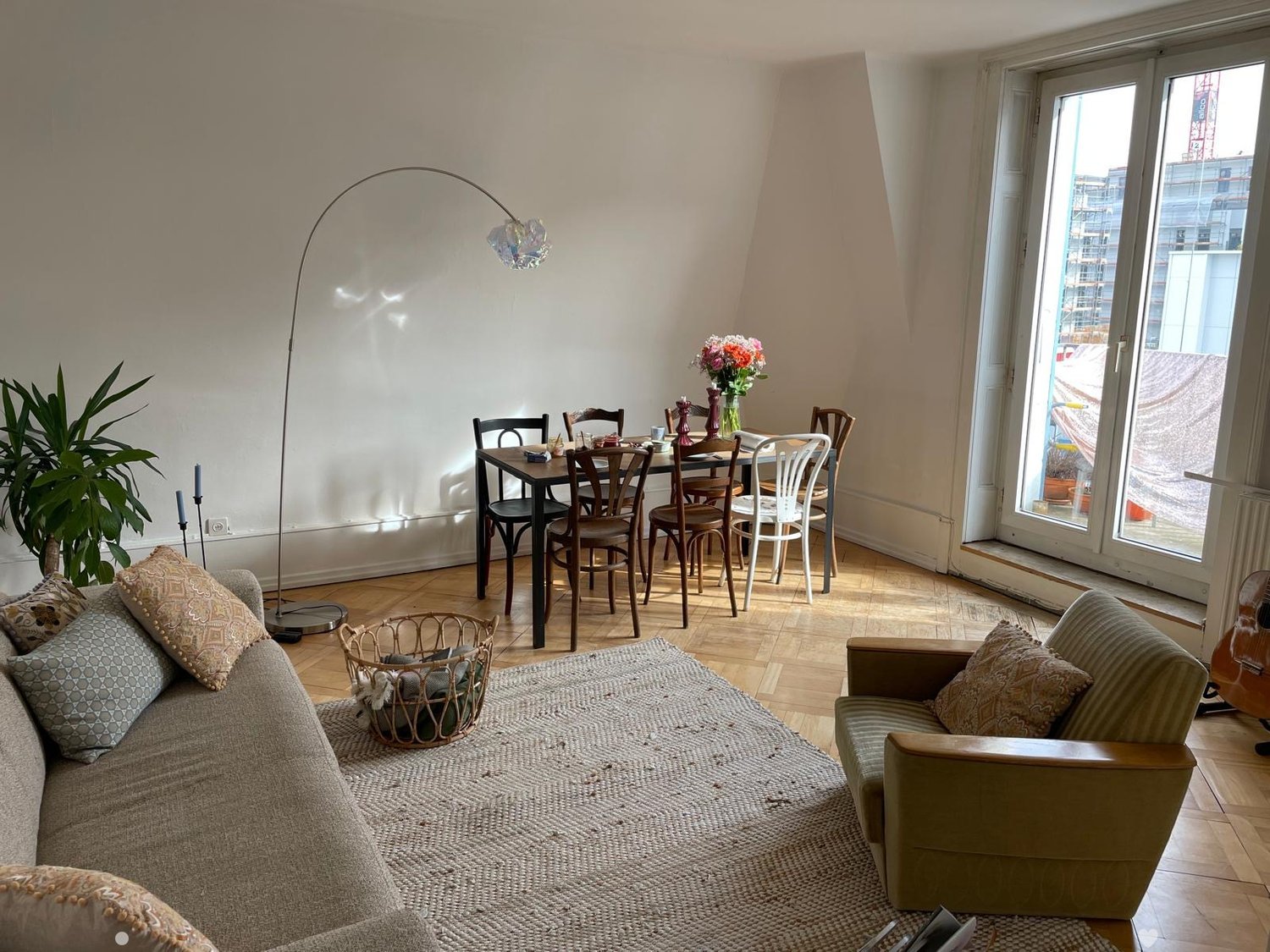 A modern, well-lit living and dining area with a grey sofa, armchair, coffee table, dining table with chairs, floor lamp, and decorative items including a plant, vase, and candles.