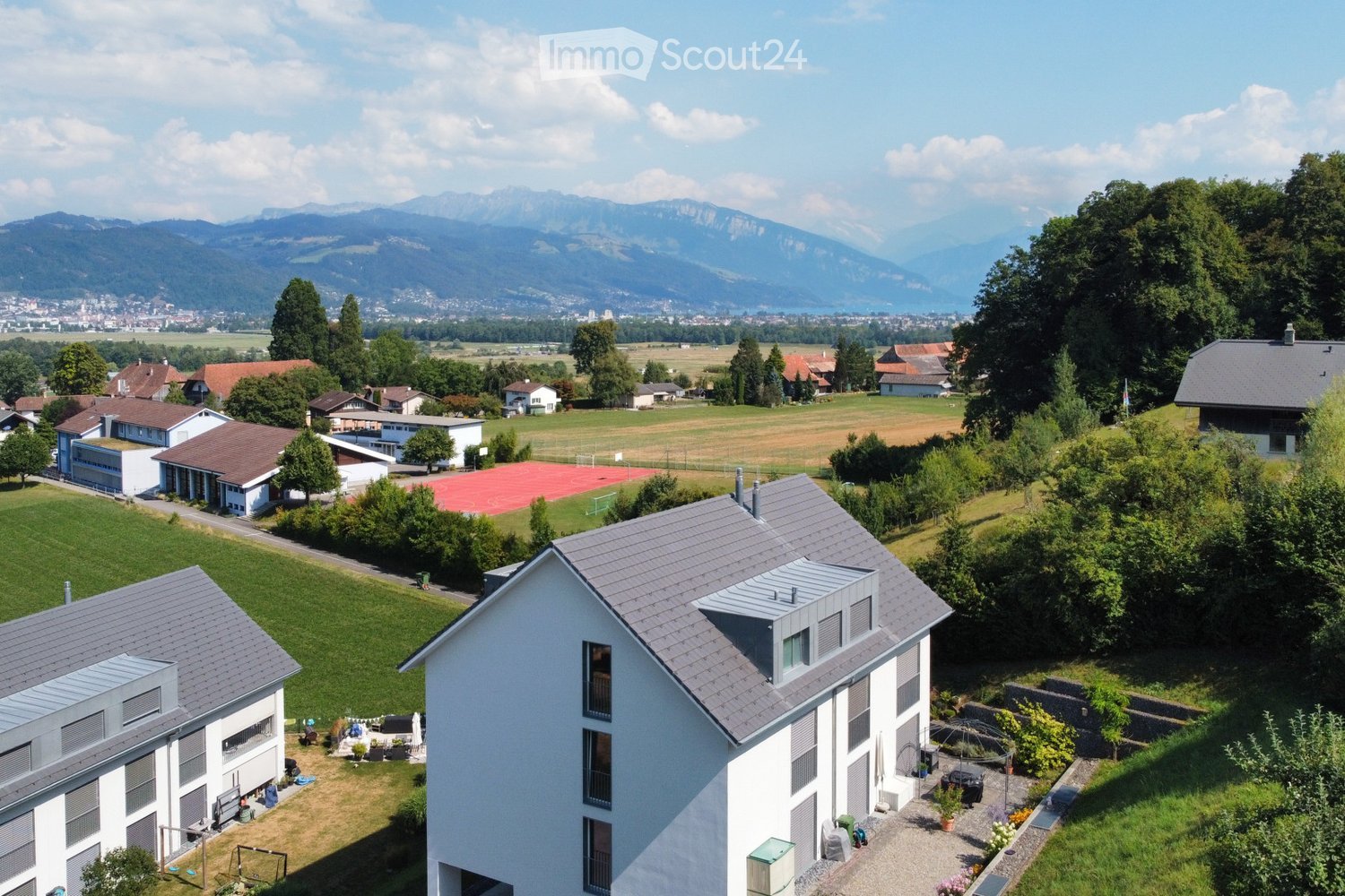 Aerial view of a residential area with multiple houses, green lawns, trees, and a distant mountain range.