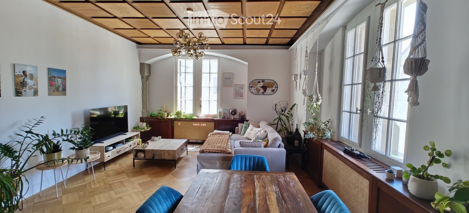 Bright living room with wooden flooring, large windows, a grey sofa, coffee table, TV, and several potted plants. Features a wooden dining table with blue chairs and a chandelier hanging from the ceiling.
