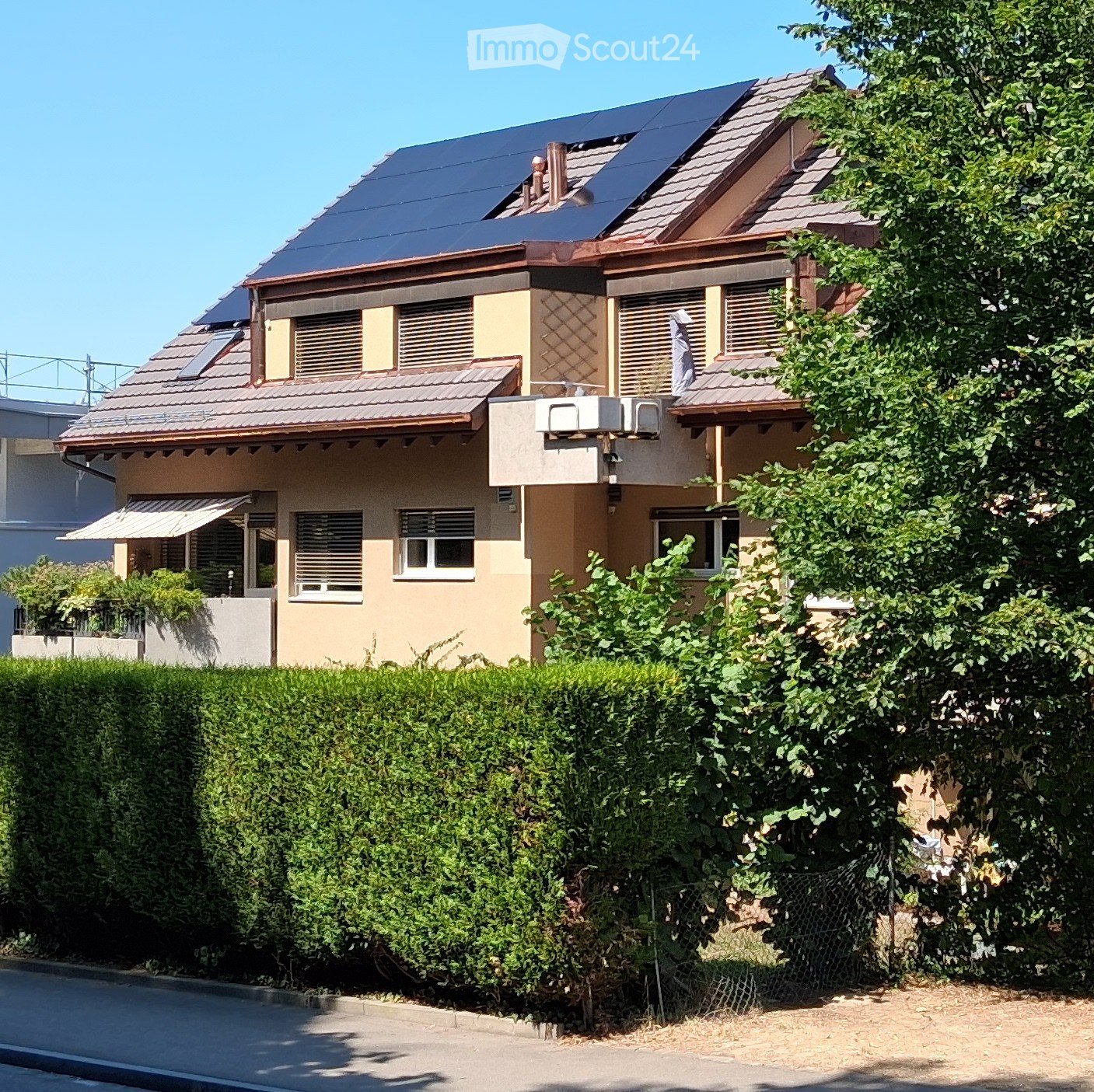 Two-story house with solar panels on the roof, beige and brown exterior, multiple windows, and a well-maintained hedge in the front yard.