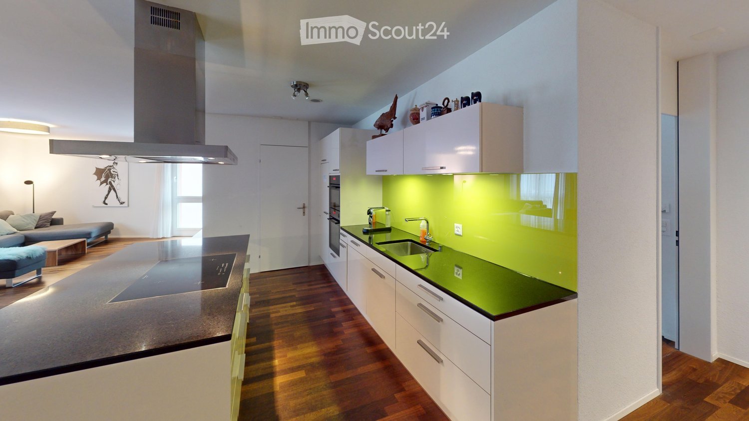 Modern kitchen and living area with white cabinets, green backsplash, wooden flooring, and integrated appliances. Features a sofa, coffee table, and a kitchen island with a built-in induction cooktop.