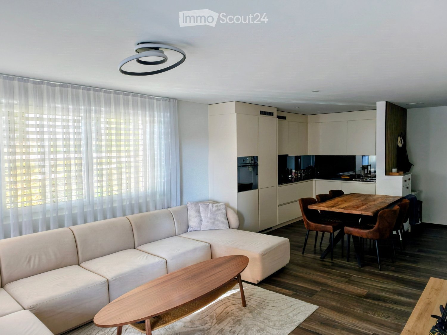 Modern living and dining area with a white sectional sofa, wooden coffee table, dining table with chairs, and a sleek kitchen area with cabinets and appliances.