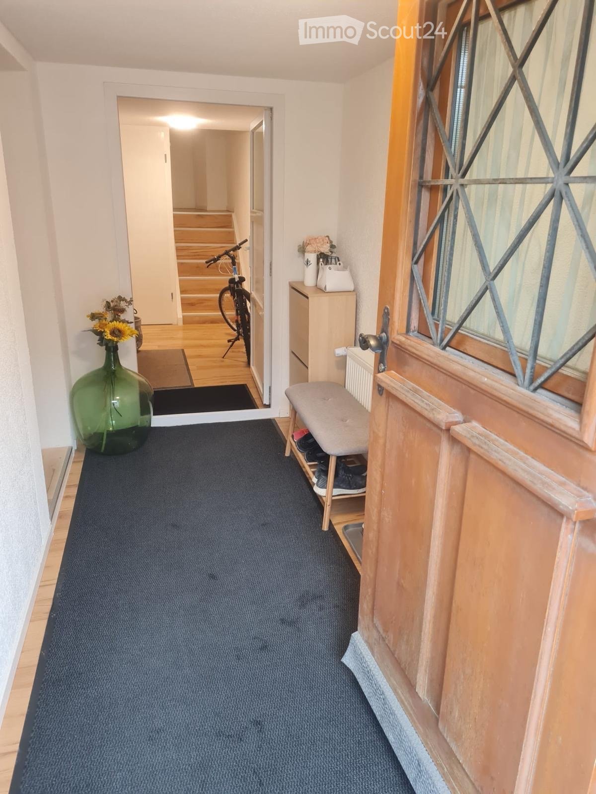 Hallway with blue carpet, wooden floor visible in adjacent area, small bench with shoes, bicycle, and decorative items including vases and flowers.