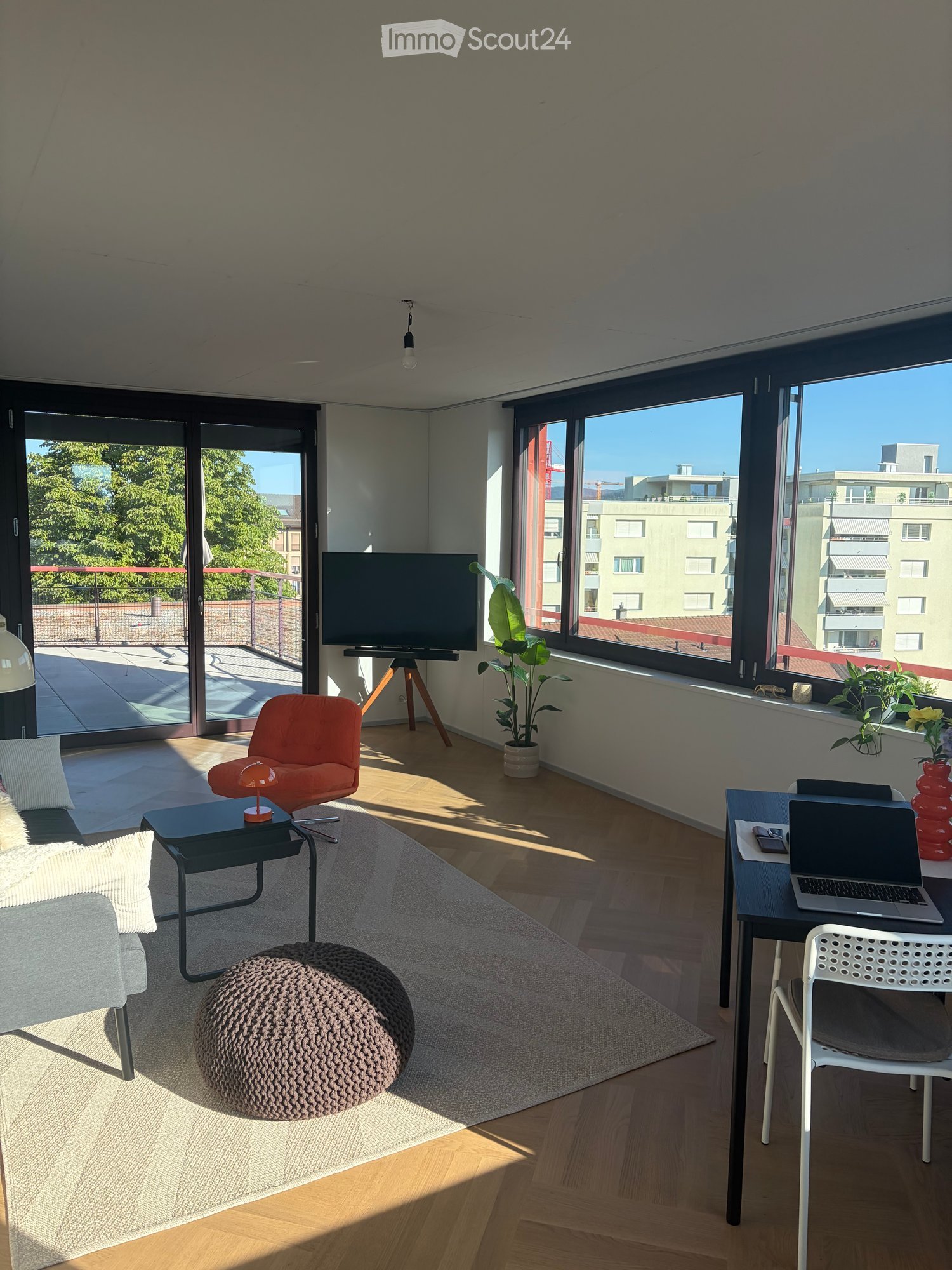 Modern living room with large windows, a balcony, a grey sofa, a round coffee table, a colorful chair, a laptop on a desk, and a television on a stand.