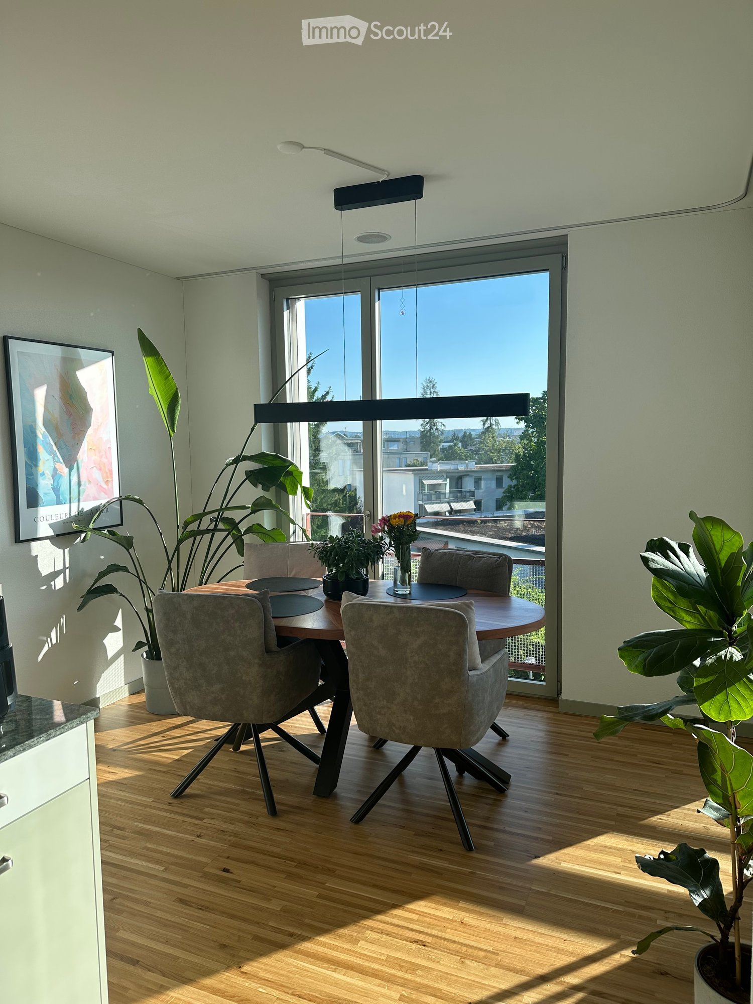 Modern dining area with wooden floor, large window offering a view, round wooden dining table with four grey upholstered chairs, potted plants, and a vase with flowers on the table.