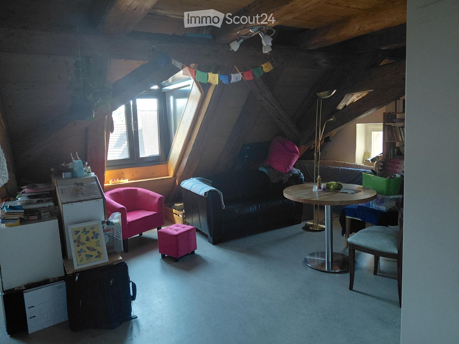 Cozy attic living space with exposed wooden beams, skylight windows, a sofa, a pink armchair, a round table with chairs, and a small pink ottoman.