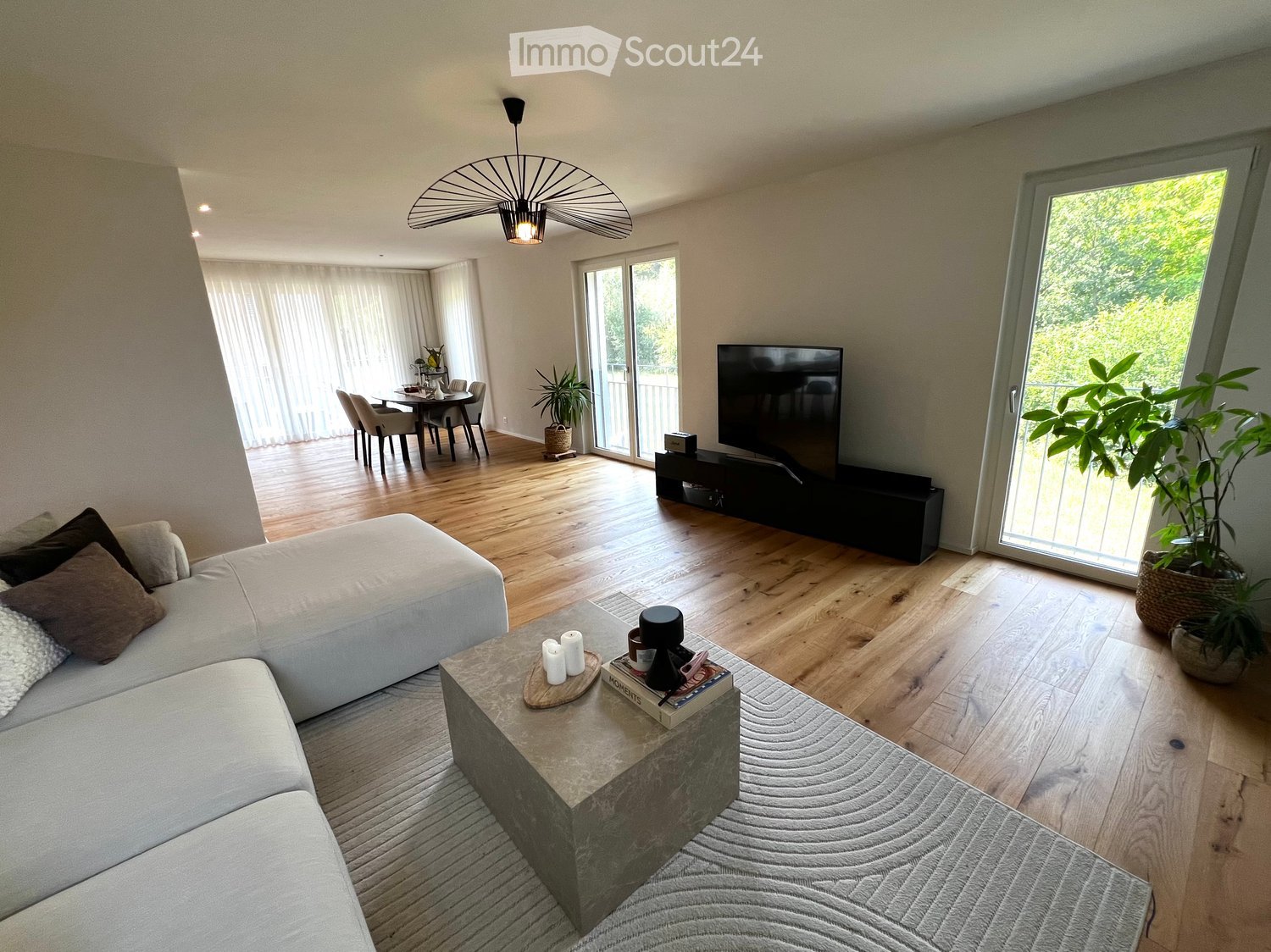 Modern living room with a white sectional sofa, coffee table, TV, dining area, and large windows with sheer curtains.