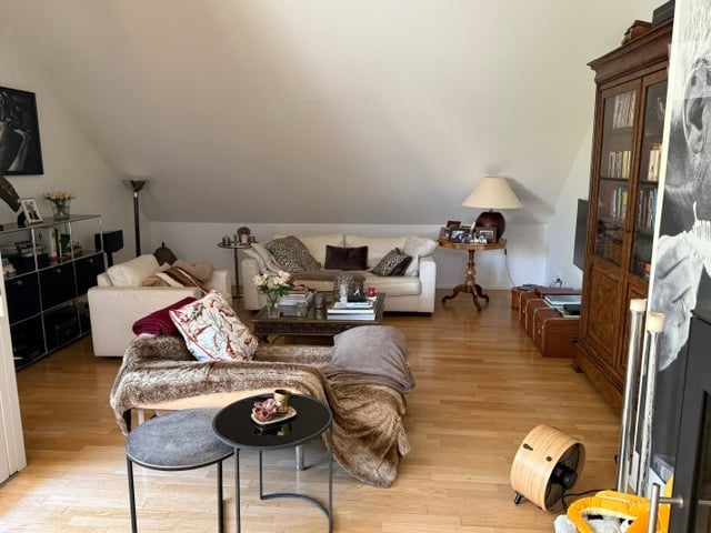 Bright living room with wooden flooring, white walls, and slanted ceiling. Features a white sectional sofa, coffee table with books and decorative items, side table with lamp, and a bookshelf with books and decorative objects.