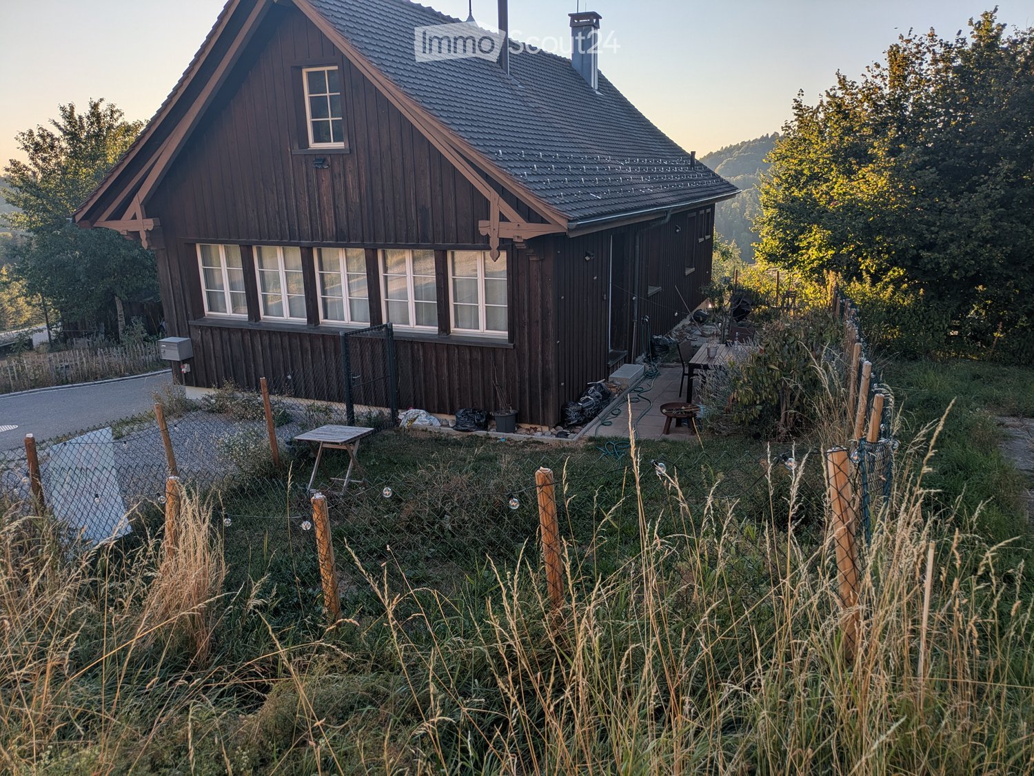 A two-story wooden house with a pitched roof, multiple windows, and a chimney, surrounded by a grassy area with a small fence and outdoor furniture.