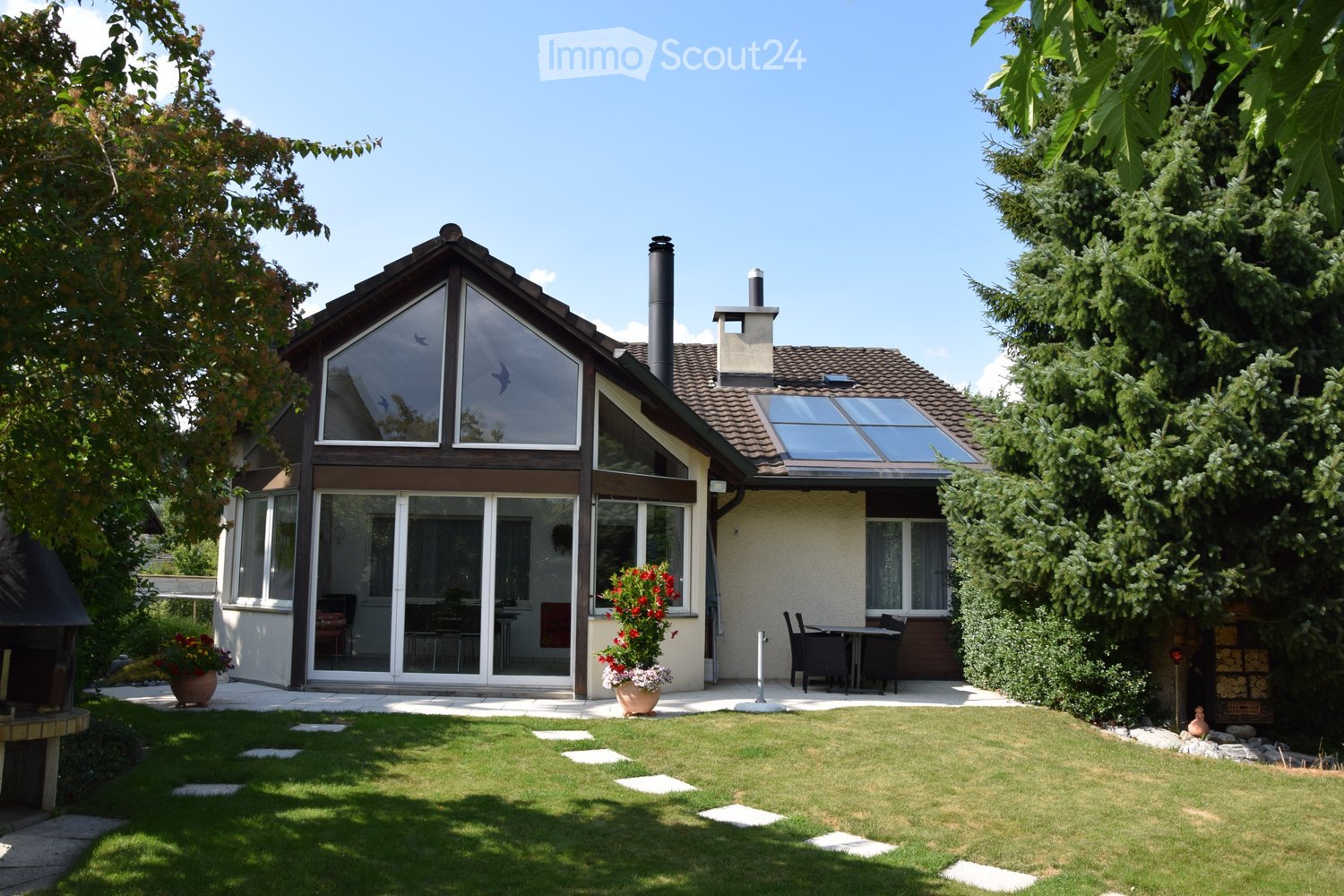 Two-story house with a modern design, large windows, and a well-maintained garden with a patio and outdoor furniture.