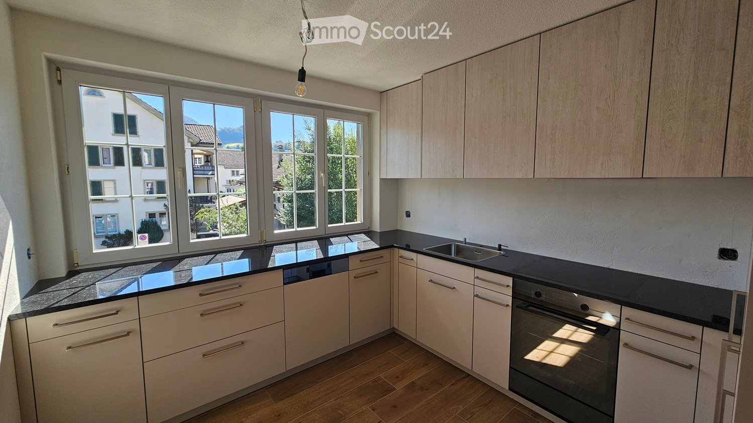 Modern kitchen with light wood cabinets, black countertops, and large windows overlooking a residential area.