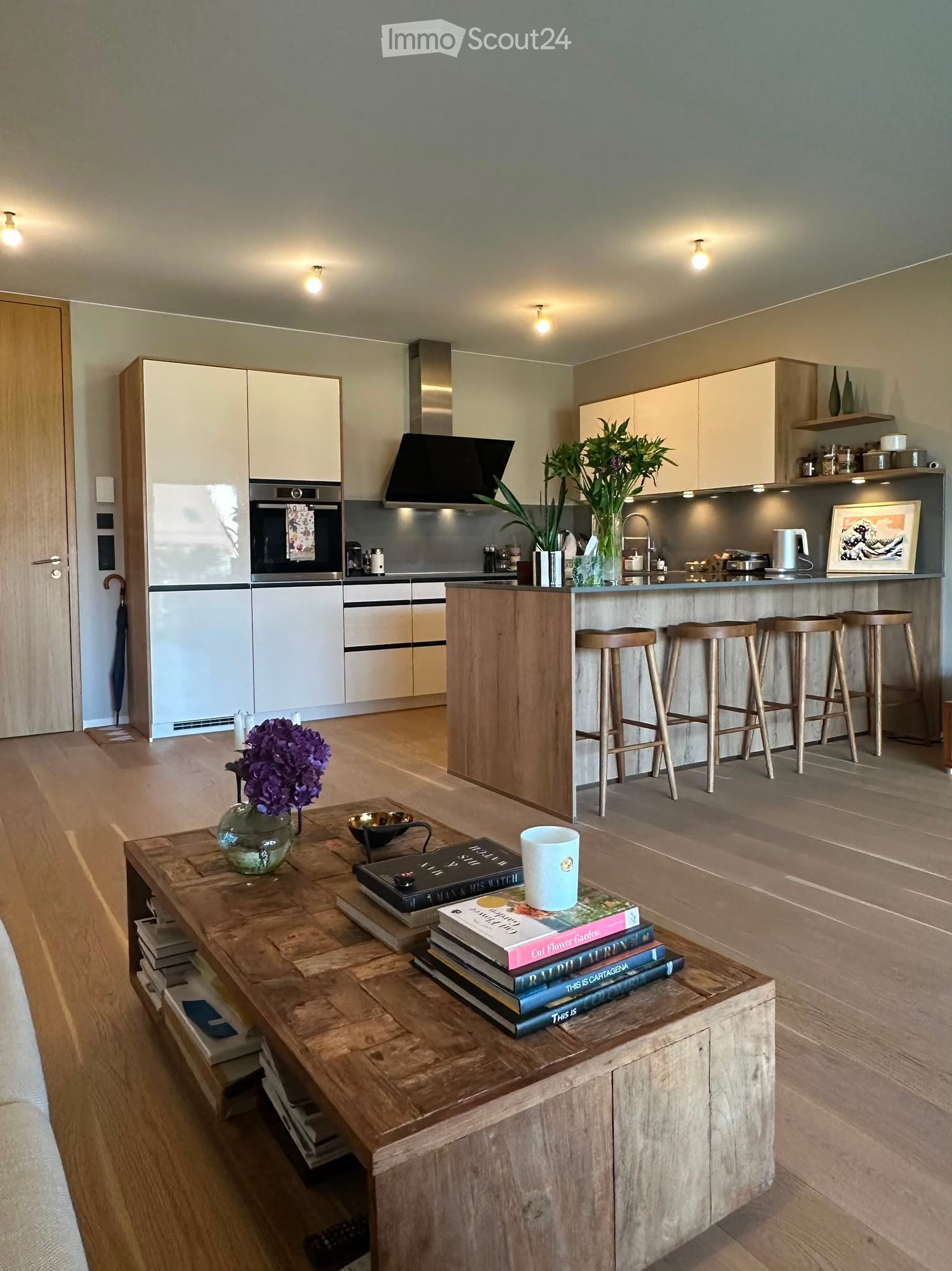 Modern kitchen and living area with light-colored cabinets, a central island with bar stools, a coffee table with books and a cup, and a vase with flowers. The kitchen features stainless steel appliances, including a refrigerator, oven, and sink.