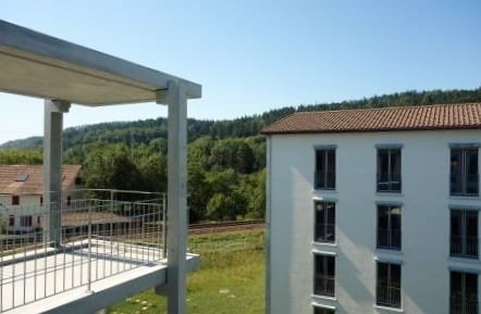 Modern multi-story building with white facade and multiple windows, featuring a balcony with metal railing and a scenic view of greenery and hills.