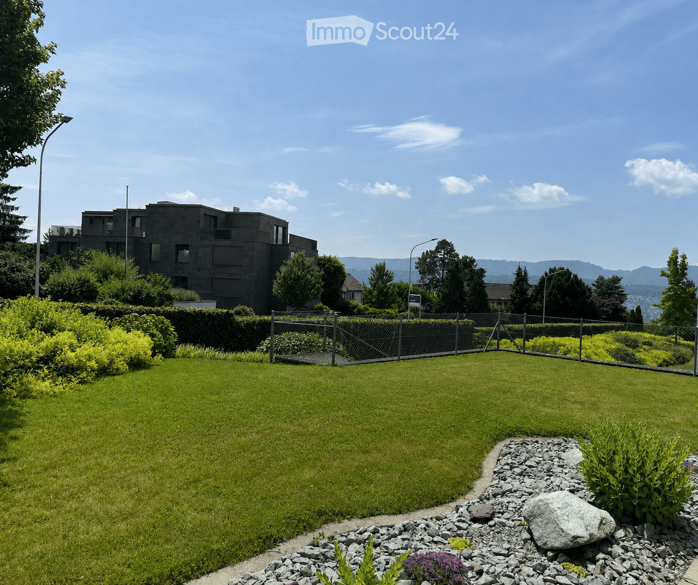 Well-maintained garden with a large lawn, rock border, and surrounding greenery. A modern building with balconies is visible in the background.