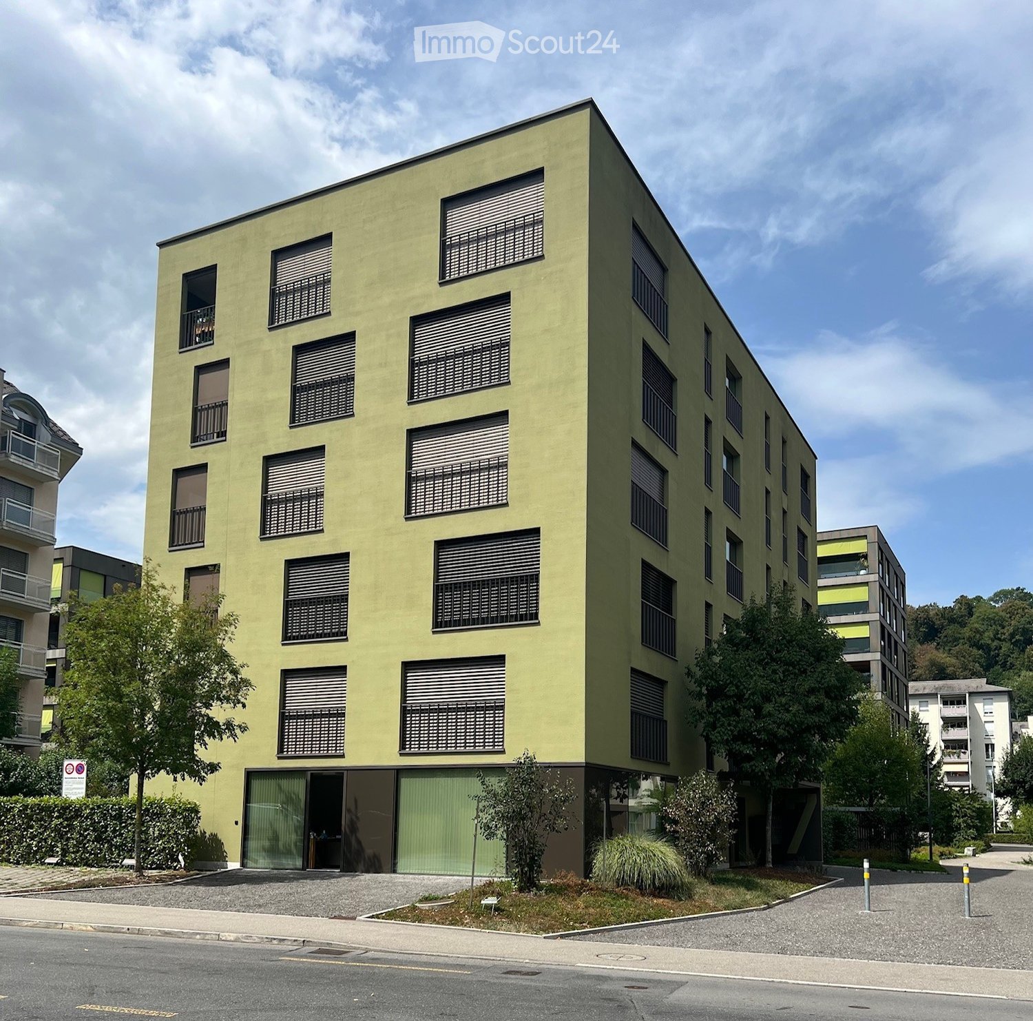 Modern multi-story building with green facade, large windows, and balconies. The building has a contemporary design with a flat roof and is surrounded by trees and greenery.