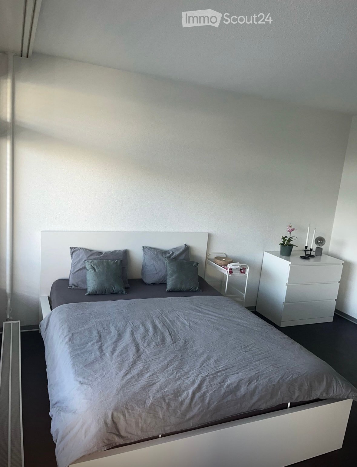 A modern bedroom with a large bed, grey bedding, and a white headboard. A white nightstand holds a small plant and candles. A small white shelf with drawers is visible next to the bed.