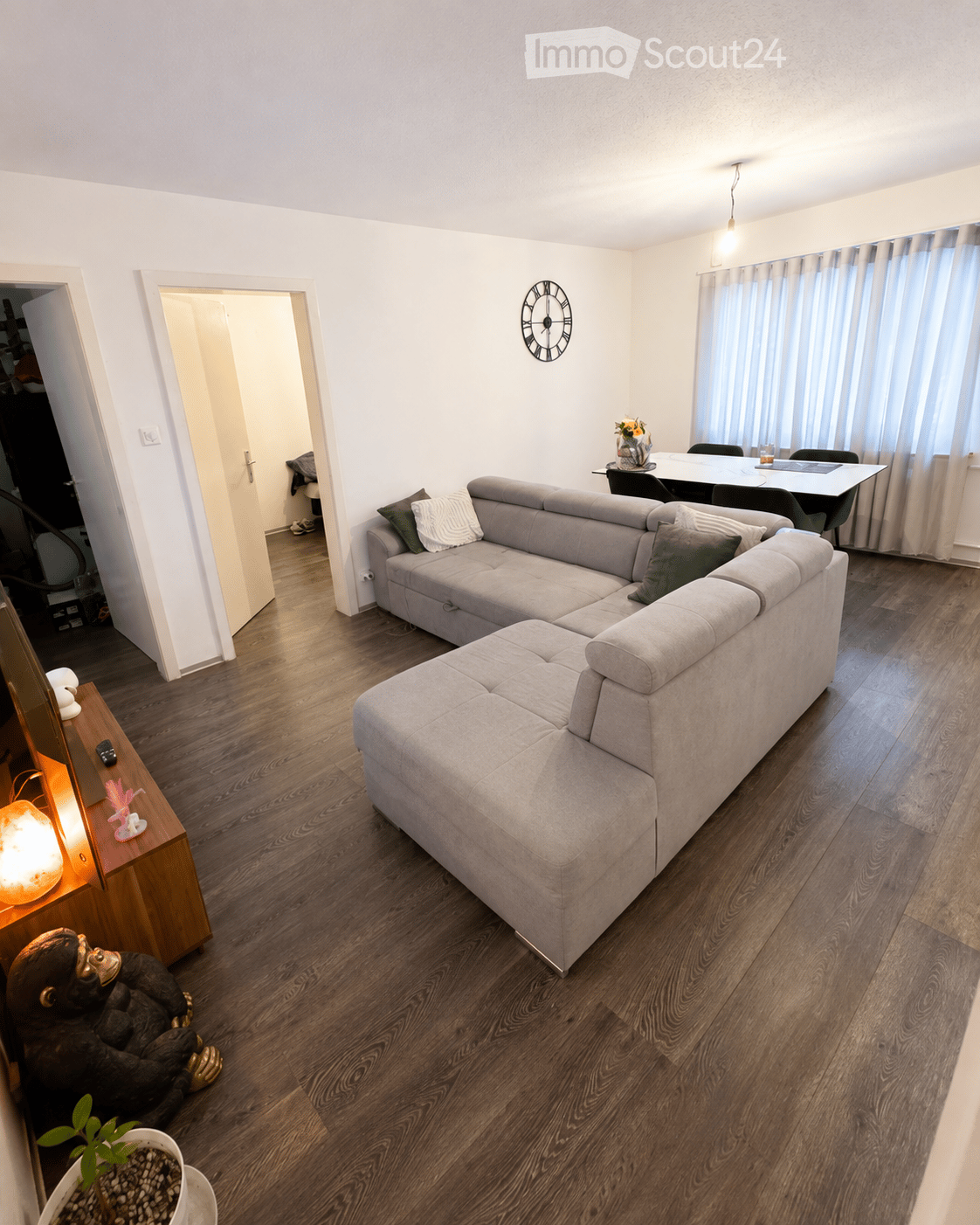 Modern living room with a grey sectional sofa, wooden coffee table, dining area with chairs and table, wall-mounted clock, and decorative items including a plant and a lamp.