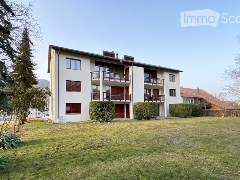 Multi-story residential building with balconies, surrounded by a lawn and trees.