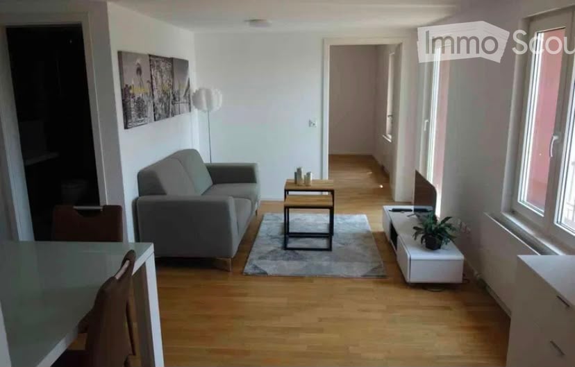 Modern living room with light wooden flooring, grey sofa, coffee table, small side table, and a plant on a white console. The room features large windows and a hallway leading to another area.
