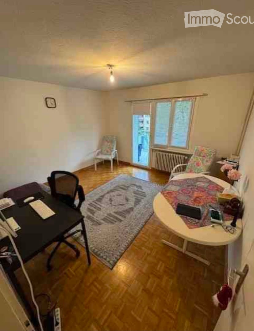 A furnished room with wooden flooring, a large rug, a desk with a laptop, a chair, and a small table with a floral-patterned chair.