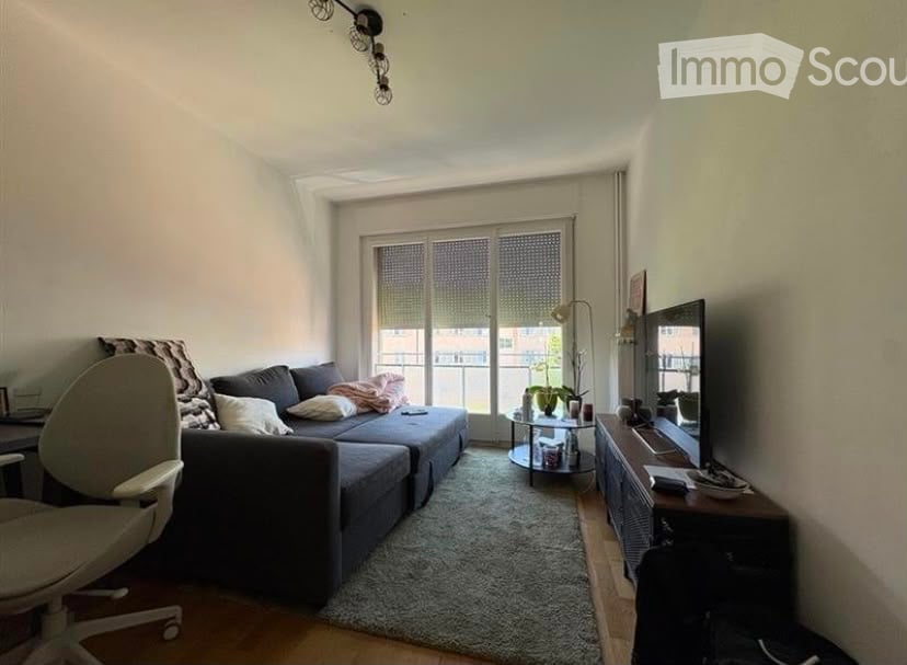 Modern living room with a grey sectional sofa, coffee table, TV stand, floor lamp, and a small round table near the balcony. The room features large windows with blinds and a cozy rug on the floor.