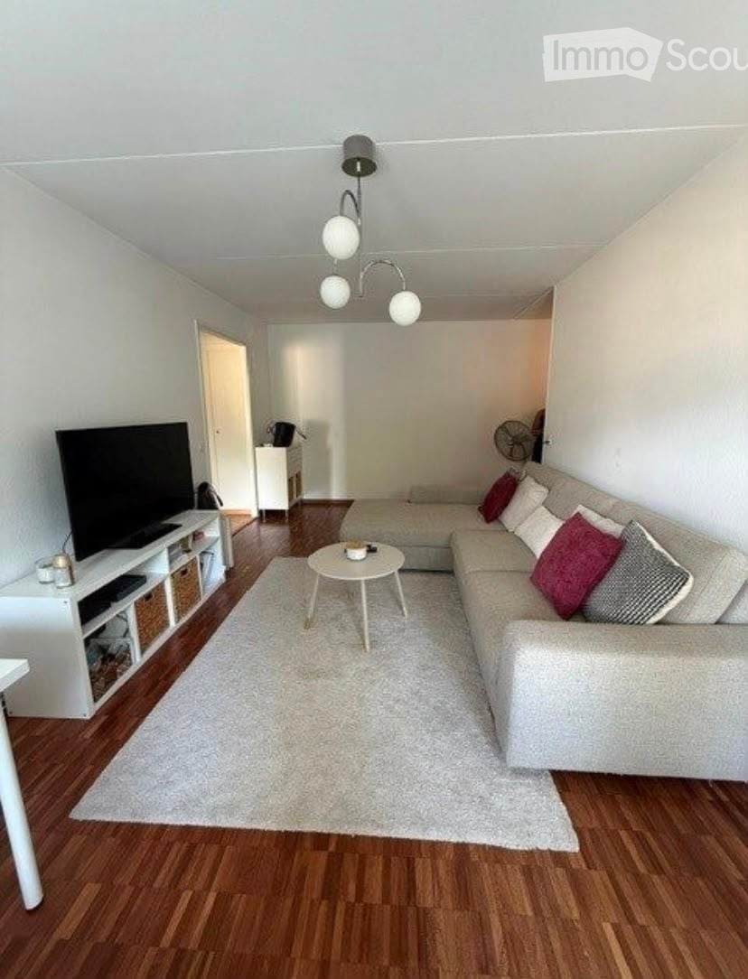 Modern living room with light-colored sofa, coffee table, TV, side table, and fan. Wooden flooring and white walls.