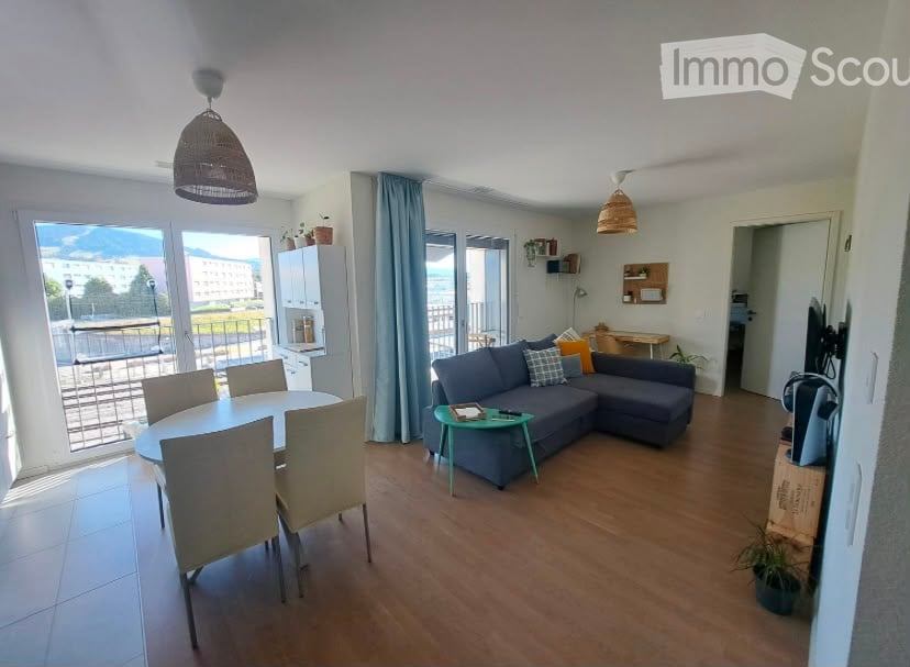Open-plan living and dining area with wooden flooring, large windows, and a balcony. Features a grey sectional sofa, coffee table, dining table with chairs, and pendant lights.