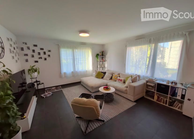 Modern living room with a large sectional sofa, coffee table, armchair, bookshelf, and decorative plants. The room features large windows with white curtains and a dark wooden floor.