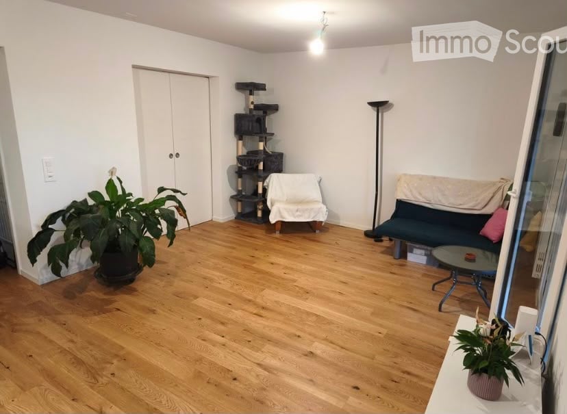 A small, well-lit living room with wooden flooring, a sofa, a coffee table, a cat tree, and a potted plant near the window.