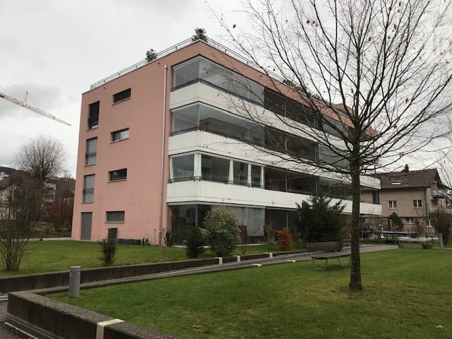 Modern multi-story building with pink facade and large glass windows, surrounded by a grassy area with trees and benches.