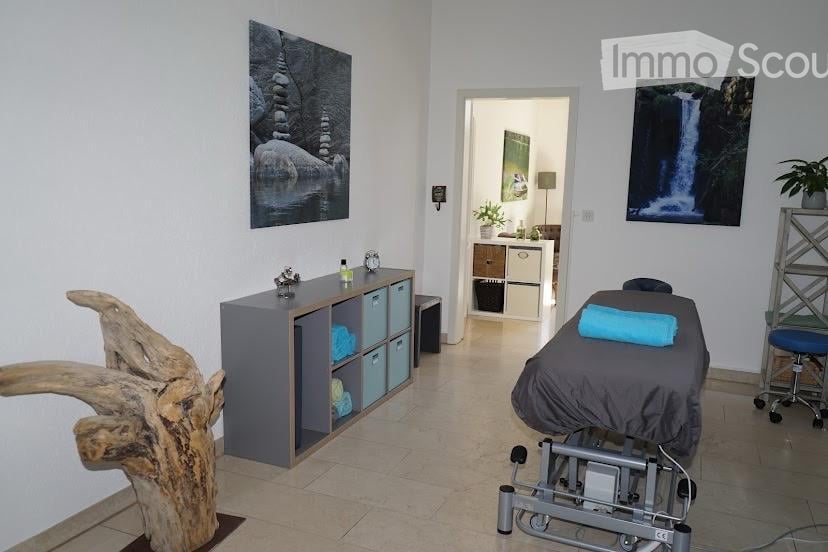 A well-lit, modern room with a massage table, storage cabinet, decorative wooden sculpture, picture frames, and a small plant. The space is clean and organized, suitable for relaxation or therapy.