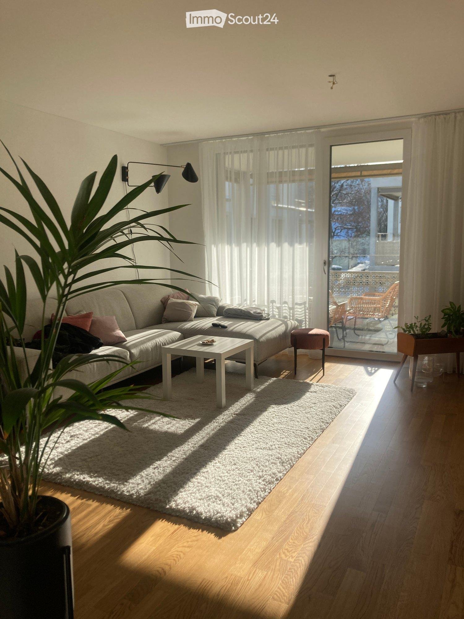 Bright living room with wooden flooring, white sofa, coffee table, wall lamp, and large window with sheer curtains leading to a balcony.
