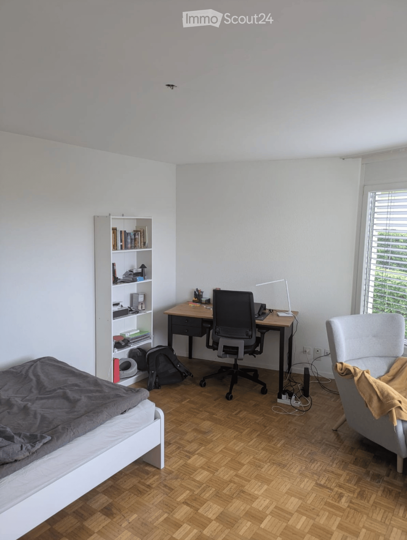 A modern, multi-functional room with a bed, desk, and seating area. Features include a wooden floor, white walls, a large window with blinds, and a bookshelf with books and storage items.