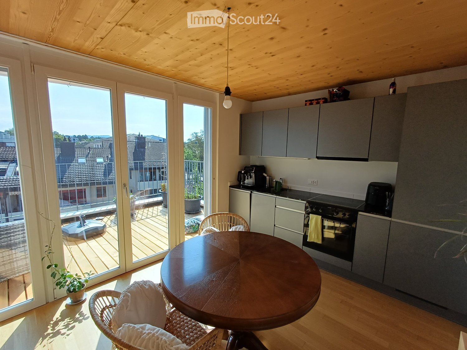 Modern kitchen with grey cabinets, a round wooden dining table, wicker chairs, and a view of a balcony with outdoor furniture.