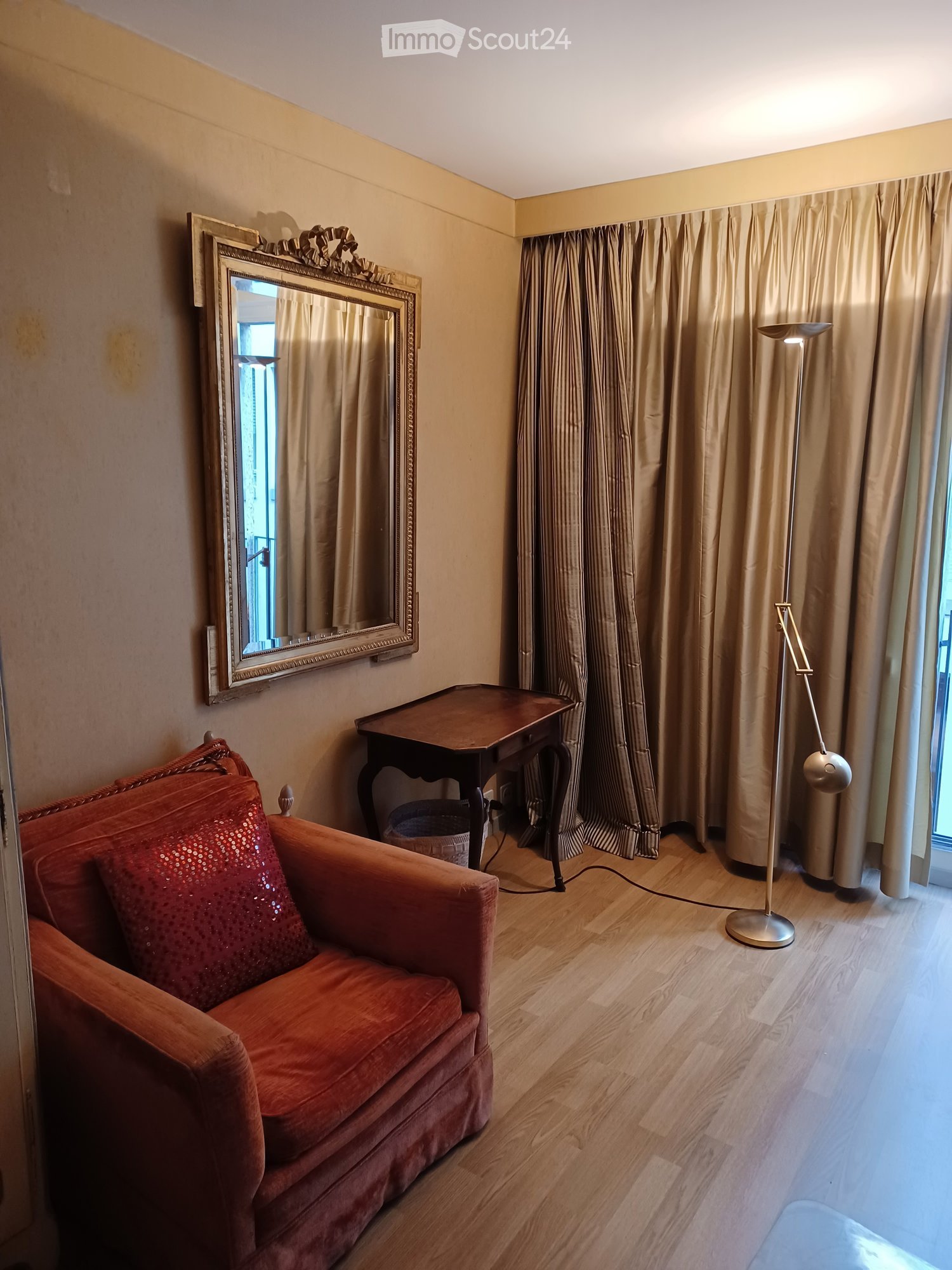 A cozy living room with a red sofa, wooden table, large mirror, and floor lamp. Beige curtains cover the windows, and the floor is made of light wood.