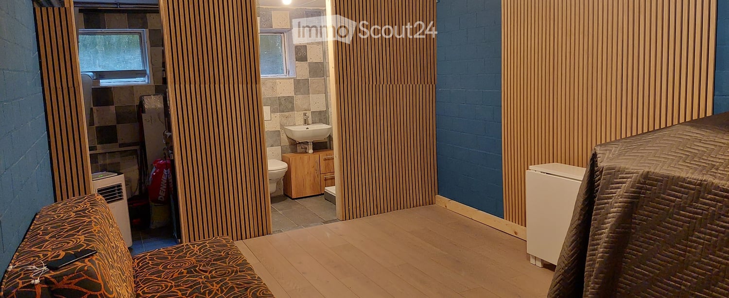 A small, furnished room with a sofa, a small table, a refrigerator, and an adjacent bathroom area with a sink, toilet, and wooden cabinet.