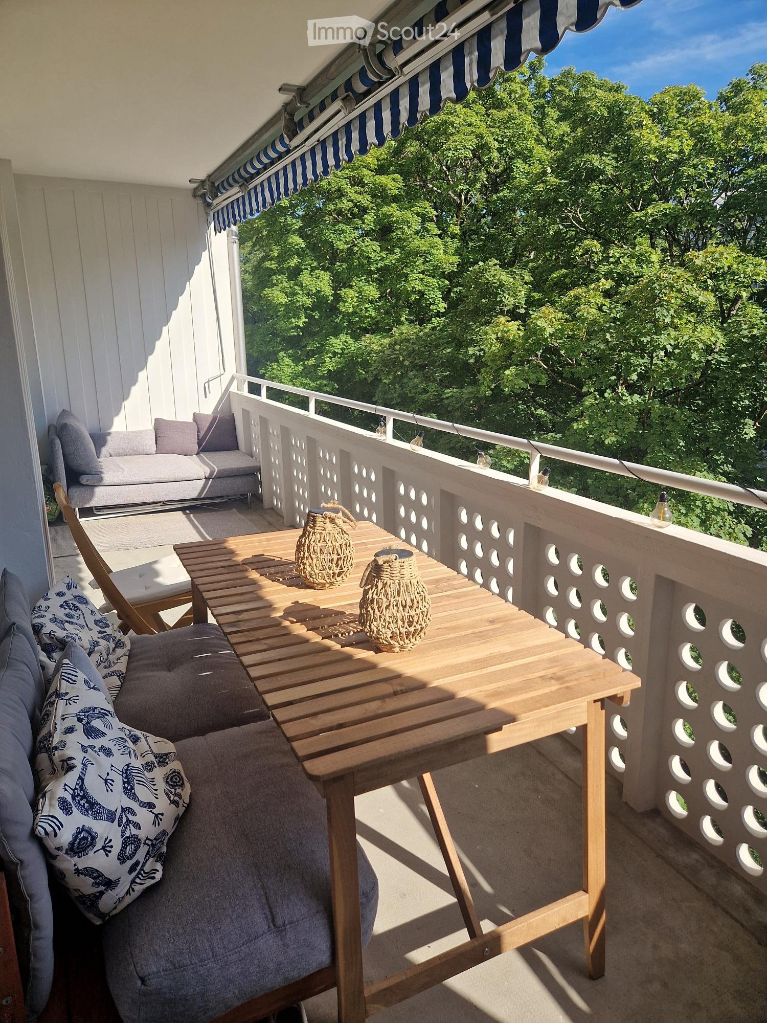 Balcony with wooden decking, sofa, table, and decorative vases, overlooking a green area.