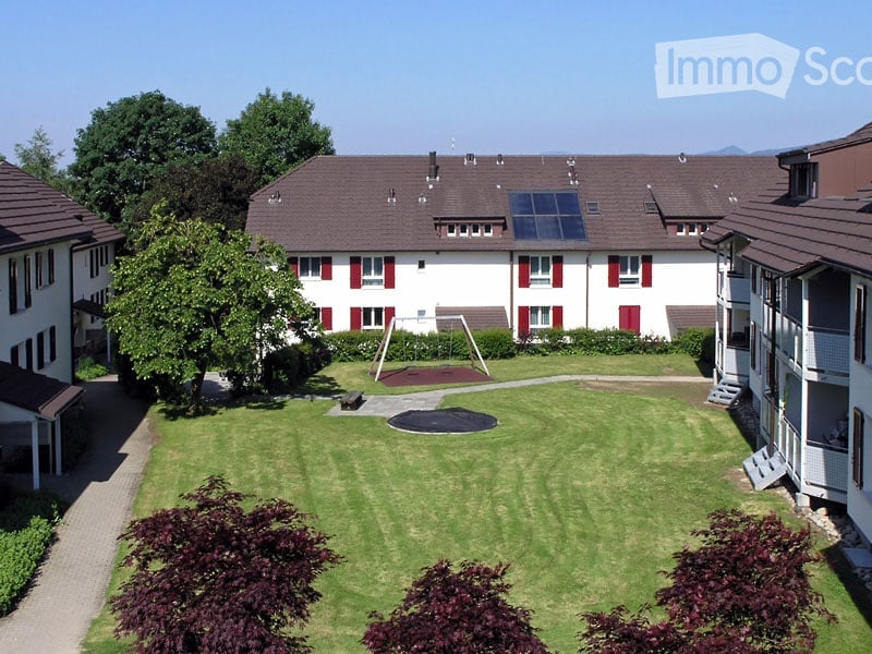 Well-maintained lawn with trimmed hedges and trees, surrounded by residential buildings with red shutters and solar panels on the roof.