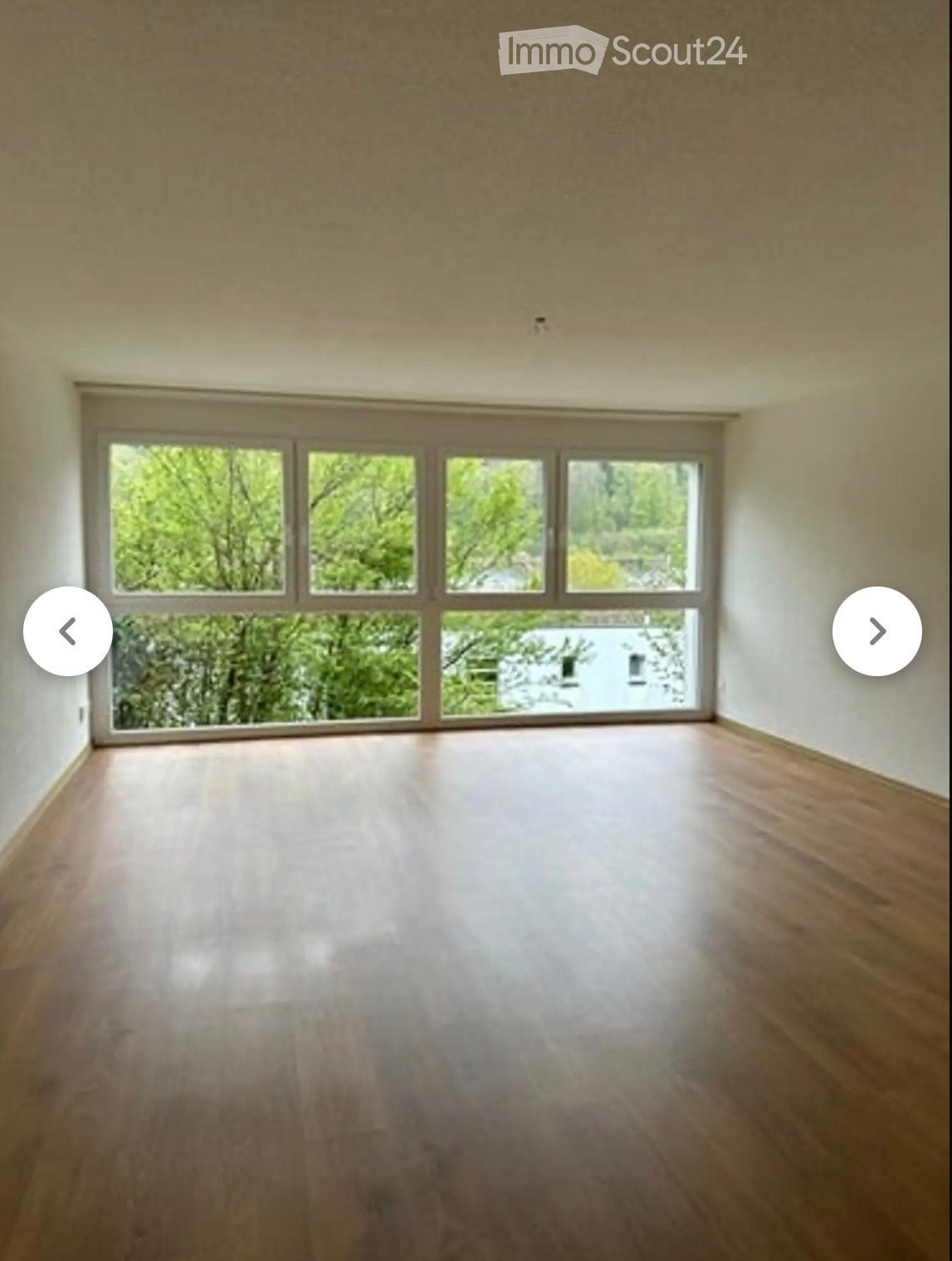 Empty room with wooden flooring and large windows showing greenery outside.
