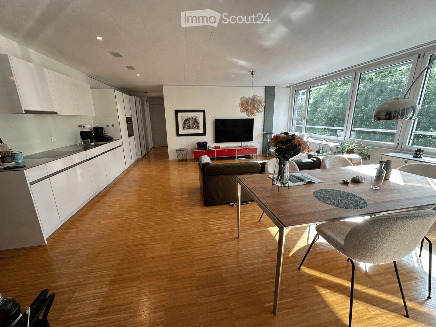 Open-plan living and dining area with modern kitchen, wooden flooring, large windows, sofa, dining table, and chairs.